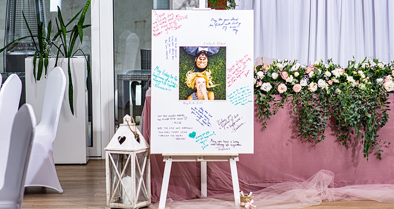Grande tela con al centro, una foto degli sposi, ed intorno, le dediche degli ospiti.  Accanto è visibile una lanterna bianca, mentre sullo sfondo c'è un tavolo decorato con fiori e tulle di colore rosa cipria
