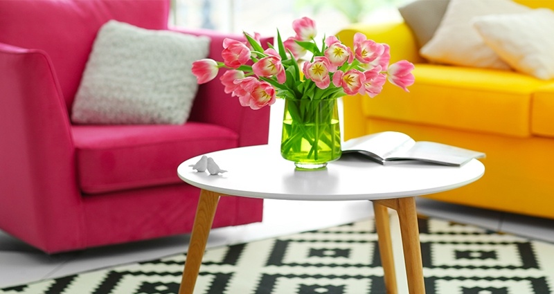 Foto che ritrae un tavolino con sopra un vaso di tulipani rosa, poggiato su un tappeto bianco e nero in salone. Sullo sfondo si vede una potrona color amaranto e un divano giallo.